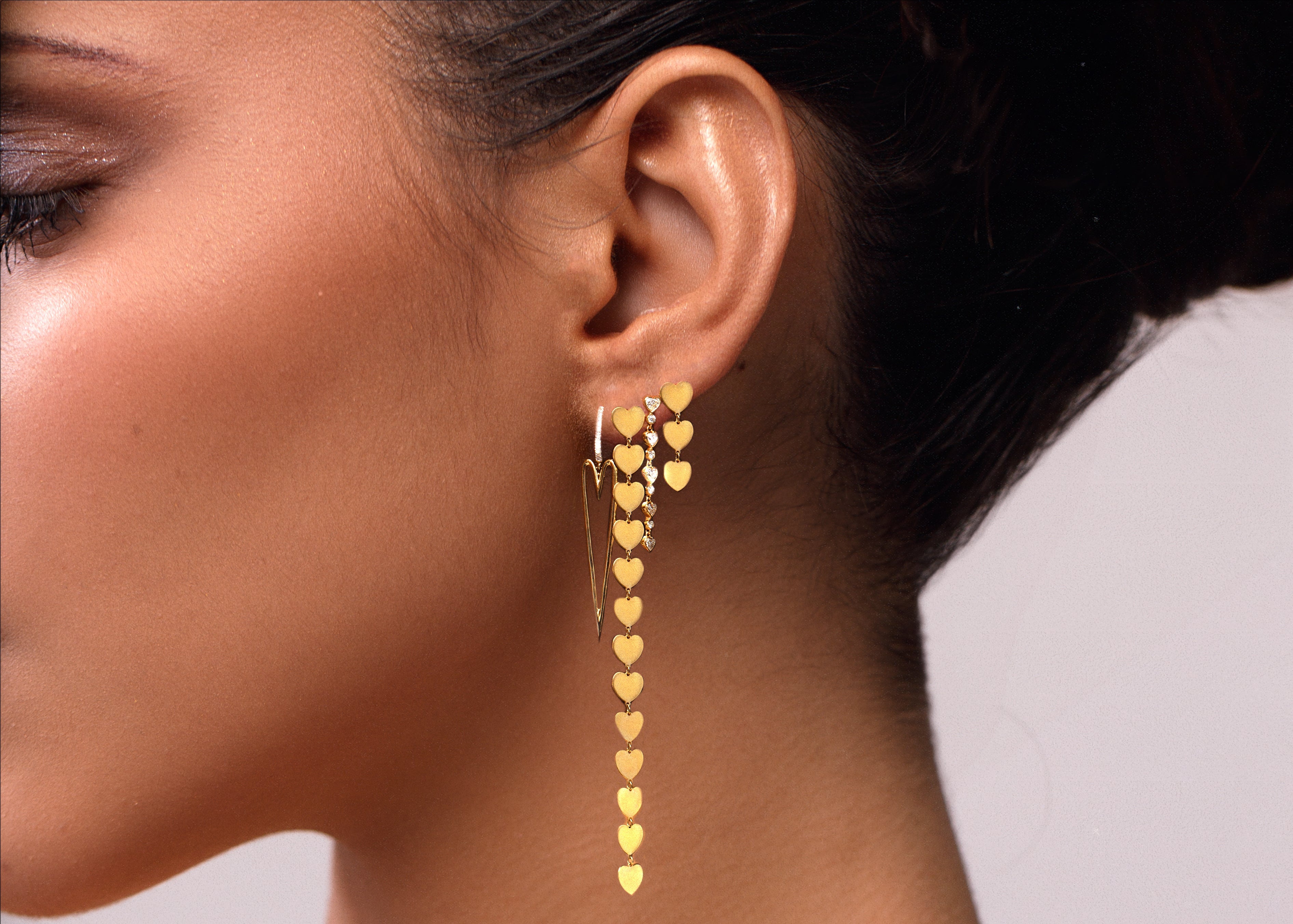 multiple designs of gold earrings with hearts shown on model's ear 