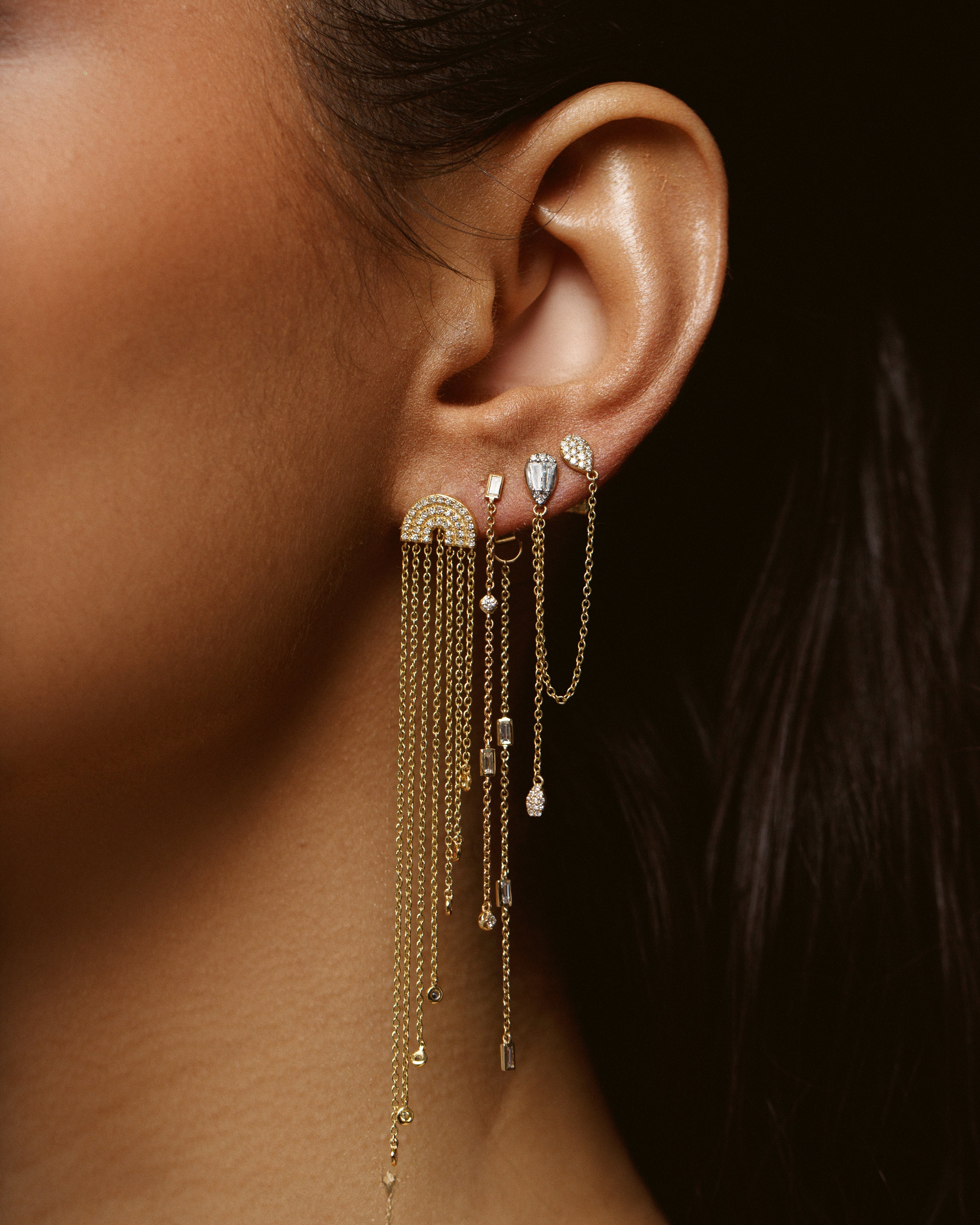 multiple gold earrings with fringe and diamond and gold chain earrings shown on model's ear